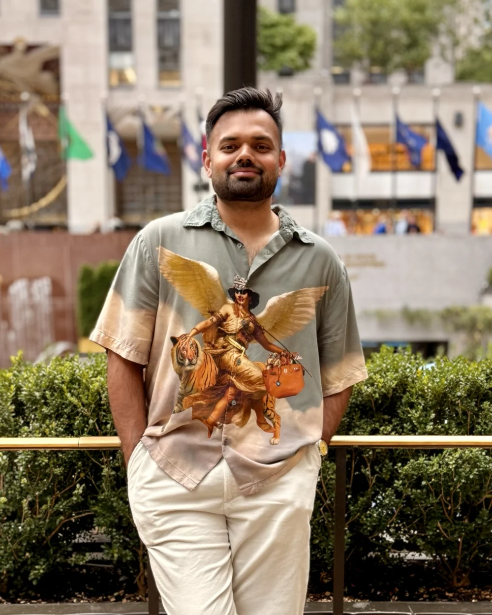 Rishabh Singh, hands in his pockets, in a printed camp-collar shirt at Rockefeller Center, with the plaza's flags and hedges behind him.